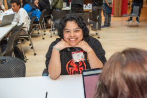 Student smiling across a table during a Mission Bit Demo Day, a day where students get to present their capstone projects, many including AI, with laptops and other students visible in the background.