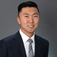 Portrait of a young Asian man in a navy suit and light grey striped tie, smiling against a neutral grey background.