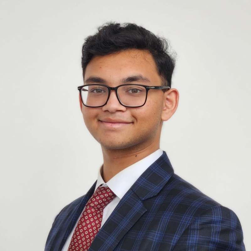 Young man in a navy blue checkered suit, white shirt, red patterned tie, glasses, smiling at the camera.