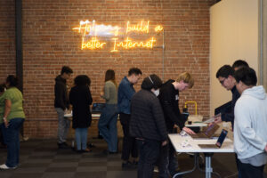 High school students present coding and technology projects during a Mission Bit Demo Day event beneath a “Helping build a better Internet” sign, showcasing hands-on computer science learning and project-based education.